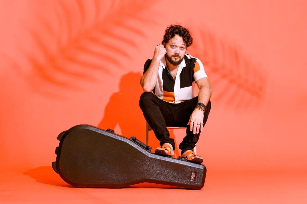 Portal Jaciara Barros – Um homem sentado em uma cadeira diante de fundo laranja, com estojo de guitarra e sombra de folha de palmeira ao fundo. – Portal Jaciara Barros