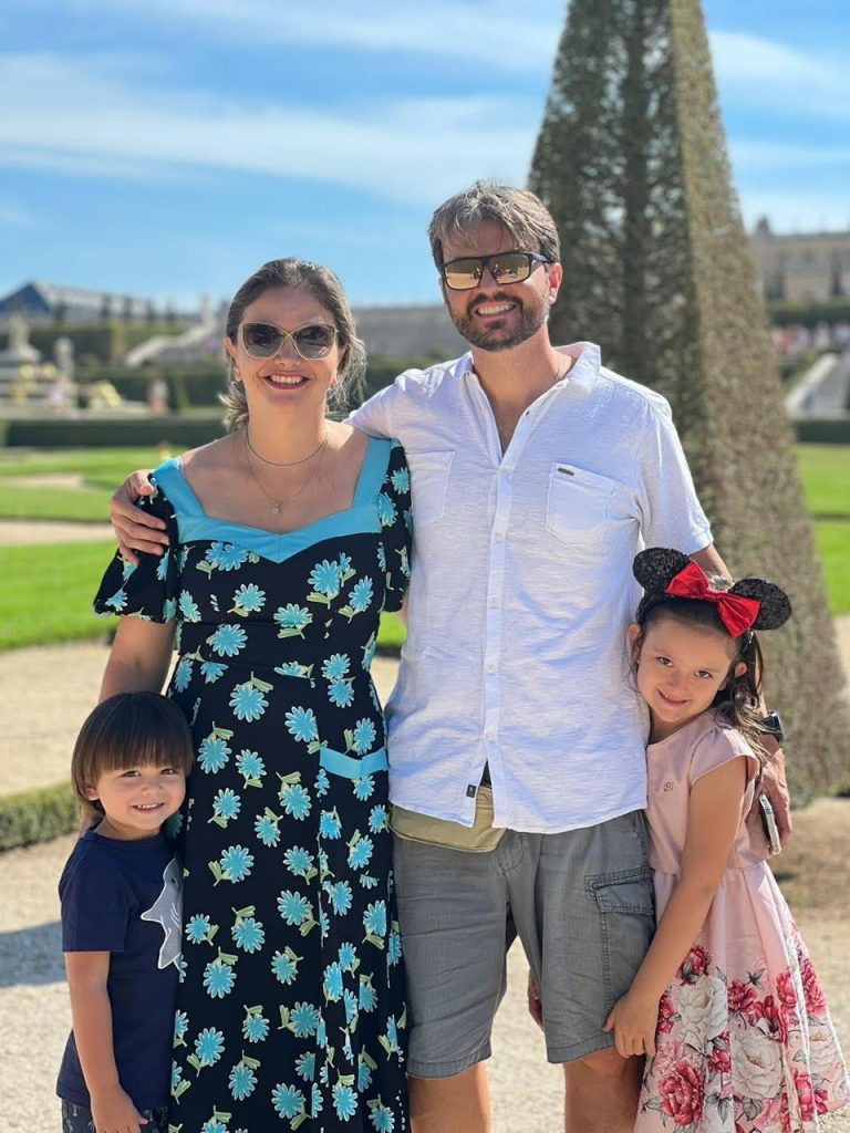 Portal Jaciara Barros – Uma família de quatro pessoas posa ao ar livre sob o sol, com jardim bem cuidado e prédio histórico ao fundo. – Portal Jaciara Barros