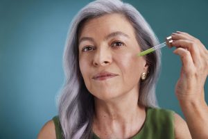 Portal Jaciara Barros – Mulher idosa de cabelos grisalhos aplica sérum verde no rosto com uma conta-gotas, em frente a um fundo azul-petróleo. – Portal Jaciara Barros