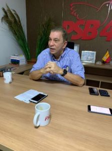 Portal Jaciara Barros – Homem sentado à mesa com papéis, canecas e telefones; ao fundo, aparecem o logotipo do PSB e livros de Amastha. – Portal Jaciara Barros