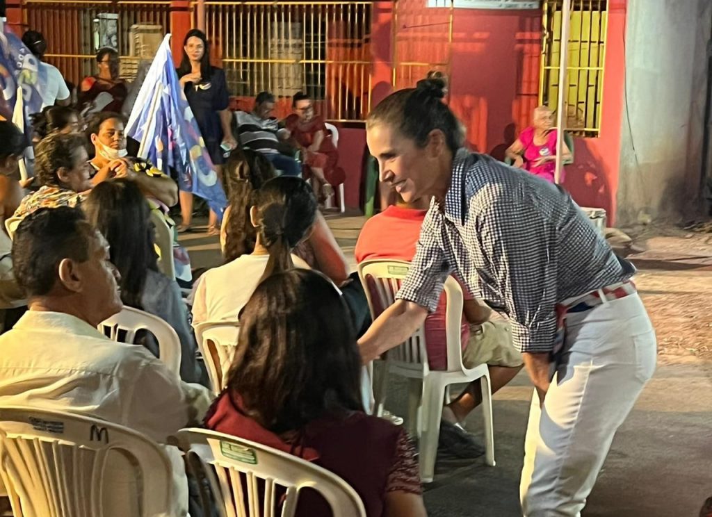 Portal Jaciara Barros – Glô Amastha, vestindo camisa xadrez, cumprimenta eleitores sentados em animado encontro comunitário ao ar livre à noite. – Portal Jaciara Barros