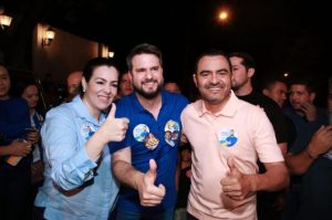 Portal Jaciara Barros – Três pessoas sorriem e fazem sinal de positivo em meio à multidão à noite, usando botons da campanha de Wanderlei Barbosa. – Portal Jaciara Barros