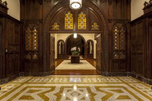 Portal Jaciara Barros – O hall de entrada da Casa Higienópolis exibe painéis de madeira trabalhada, vitrais coloridos e mesa central com flores brancas. – Portal Jaciara Barros