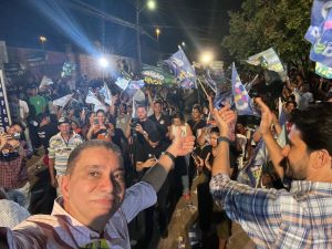 Portal Jaciara Barros – Homem faz uma selfie em evento lotado à noite, com pessoas erguendo bandeiras sob luz intensa. – Portal Jaciara Barros