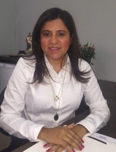 Portal Jaciara Barros – Uma mulher de cabelos longos e escuros senta-se à mesa, com camisa branca e colares, mãos cruzadas, pronta para o dia. – Portal Jaciara Barros