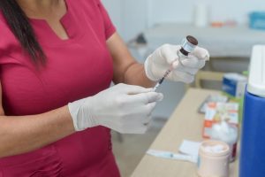 Portal Jaciara Barros – Profissional de uniforme vermelho prepara seringa para vacinação contra Covid-19 usando luvas brancas em ambiente clínico. – Portal Jaciara Barros