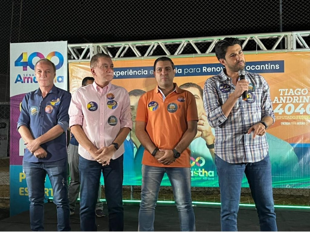Portal Jaciara Barros – Tiago Andrino discursa ao microfone ao lado de três homens em evento, com placas de campanha ao fundo. – Portal Jaciara Barros