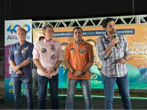 Portal Jaciara Barros – Tiago Andrino discursa ao microfone ao lado de três homens em evento, com placas de campanha ao fundo. – Portal Jaciara Barros