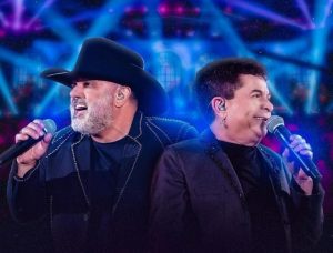 Portal Jaciara Barros – Dois homens de jaquetas escuras cantam ao microfone no palco, iluminados por luzes azul e roxa. – Portal Jaciara Barros