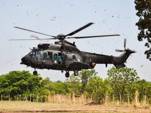 Portal Jaciara Barros – Um helicóptero preto paira próximo ao solo em Tocantins, levantando detritos perto de árvores e uma cerca. – Portal Jaciara Barros