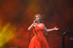 Portal Jaciara Barros – Vanessa da Mata se apresenta no palco com vestido vermelho, diante do público, sob luzes amarelas e alaranjadas ao fundo. – Portal Jaciara Barros