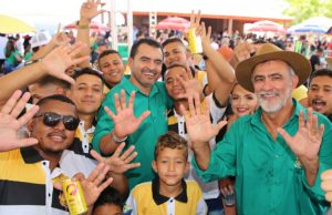 Portal Jaciara Barros – Em São Miguel, um grupo de pessoas sorri e ergue as mãos durante evento ao ar livre de assistência humanitária. – Portal Jaciara Barros