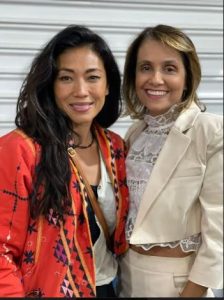 Portal Jaciara Barros – Duas mulheres, uma de jaqueta vermelha estampada e outra de roupa creme, sorriem em evento do Grupo Sallo projetos sociais. – Portal Jaciara Barros