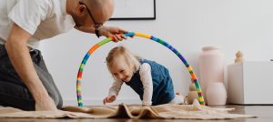Portal Jaciara Barros – Um adulto segura um arco colorido enquanto uma criança engatinha por baixo dele, promovendo educação em ambiente lúdico. – Portal Jaciara Barros