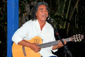 Portal Jaciara Barros – Homem de cabelos cacheados canta e toca violão ao ar livre no evento Vivência Lixo ao Luxo, vestindo camisa branca. – Portal Jaciara Barros