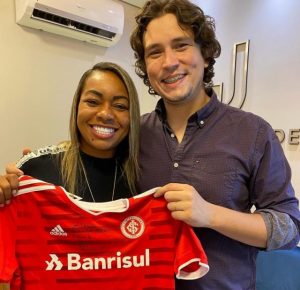 Portal Jaciara Barros – O Dr. Juliano Guedes e a jogadora Sorriso sorriem enquanto exibem, em ambiente interno, a camisa do Inter com logotipo do Banrisul. – Portal Jaciara Barros