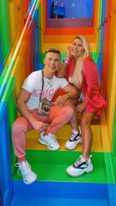 Portal Jaciara Barros – Duas pessoas sorriem sentadas em uma escadaria colorida em Brasília, vestidas com looks rosa no estilo Ken Humano e Barbie Gaúcha. – Portal Jaciara Barros