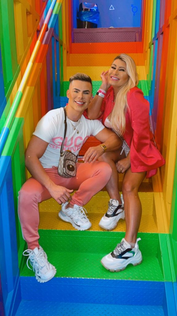 Portal Jaciara Barros – Duas pessoas sorriem sentadas em uma escadaria colorida em Brasília, vestidas com looks rosa no estilo Ken Humano e Barbie Gaúcha. – Portal Jaciara Barros