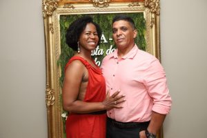 Portal Jaciara Barros – Uma mulher de vestido vermelho e um homem de camisa rosa posam lado a lado diante de uma moldura dourada ornamentada. – Portal Jaciara Barros