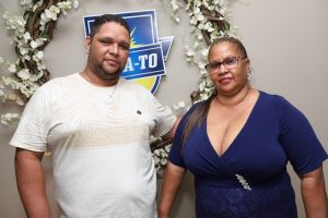 Portal Jaciara Barros – Um homem de camisa clara e uma mulher de vestido azul estão ao lado de uma guirlanda floral e de um letreiro temático soldado. – Portal Jaciara Barros