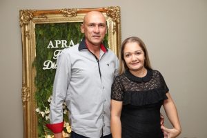 Portal Jaciara Barros – Um homem e uma mulher posam juntos em um ambiente interno, diante de um painel verde com tema soldado, letras brancas e flores. – Portal Jaciara Barros