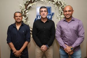 Portal Jaciara Barros – Três homens, entre eles um soldado, posam em ambiente interno diante de uma coroa de flores e uma placa com a inscrição APITO. – Portal Jaciara Barros