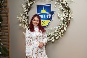 Portal Jaciara Barros – Uma mulher de cabelos cacheados ruivos posa ao lado de uma guirlanda floral e de uma placa azul com os dizeres PRA-TO. – Portal Jaciara Barros