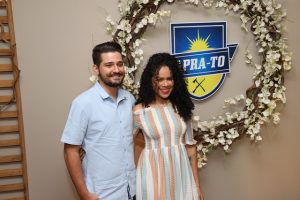 Portal Jaciara Barros – Um homem e uma mulher sorriem diante de uma coroa de flores e um emblema azul e amarelo escrito PRA-TO em homenagem a um soldado. – Portal Jaciara Barros