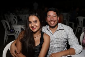 Portal Jaciara Barros – Uma mulher de vestido preto e um homem trajado como soldado sorriem juntos sentados à mesa com cadeiras plásticas brancas. – Portal Jaciara Barros