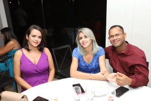 Portal Jaciara Barros – Três pessoas, entre elas um soldado, estão sentadas em uma mesa redonda com bebidas e pratos durante evento em local fechado. – Portal Jaciara Barros