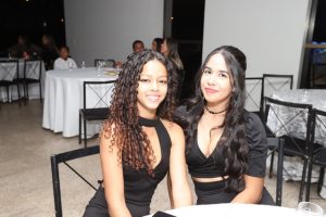 Portal Jaciara Barros – Dois jovens de vestido preto sentam-se à mesa de banquete, sorrindo com confiança e parecendo companheiras próximas. – Portal Jaciara Barros