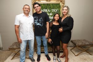 Portal Jaciara Barros – Quatro pessoas posam sorrindo ao lado de uma placa dourada decorativa e parede de musgo, com fundo inspirado em soldados. – Portal Jaciara Barros