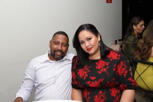 Portal Jaciara Barros – Um homem de camisa branca e uma mulher de vestido preto com flores vermelhas posam à mesa ao lado de um soldado. – Portal Jaciara Barros