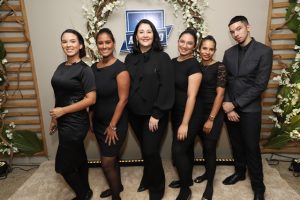 Portal Jaciara Barros – Seis pessoas em traje social preto posam para foto em grupo diante de um painel decorado, alinhadas lado a lado. – Portal Jaciara Barros