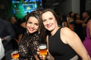 Portal Jaciara Barros – Duas mulheres sorriem e seguram copos de cerveja em evento temático soldado, com luzes coloridas ao fundo. – Portal Jaciara Barros