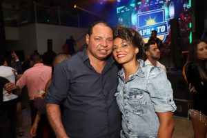 Portal Jaciara Barros – Um homem e uma mulher sorriem juntos durante evento interno, com palco colorido de tema soldado ao fundo. – Portal Jaciara Barros