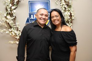 Portal Jaciara Barros – Duas pessoas sorriem lado a lado diante de uma coroa de flores e de uma placa azul em homenagem a um soldado. – Portal Jaciara Barros