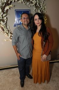 Portal Jaciara Barros – Um homem de camisa cinza e uma mulher de mostarda sorriem juntos diante de um vibrante arranjo floral. – Portal Jaciara Barros