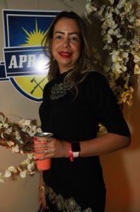 Portal Jaciara Barros – Uma mulher de preto posa com uma taça diante de um fundo floral e placa da APRA, demonstrando postura confiante. – Portal Jaciara Barros