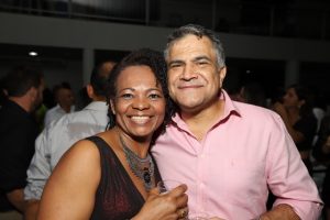 Portal Jaciara Barros – Uma mulher e um homem, este em uniforme de soldado, posam sorrindo juntos em evento interno com outras pessoas ao fundo. – Portal Jaciara Barros