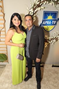 Portal Jaciara Barros – Uma mulher de vestido verde e um homem de terno escuro posam junto a um arranjo floral e placa da APRA-TO com elegância. – Portal Jaciara Barros