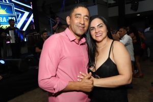 Portal Jaciara Barros – Um homem de camisa rosa e uma mulher de vestido preto posam sorrindo em evento fechado, com convidados ao fundo. – Portal Jaciara Barros