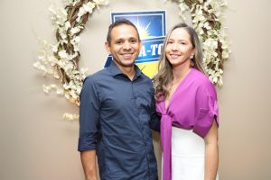 Portal Jaciara Barros – Duas pessoas sorriem em frente a uma parede decorada com guirlanda de flores e uma placa soldado azul e amarela ao fundo. – Portal Jaciara Barros