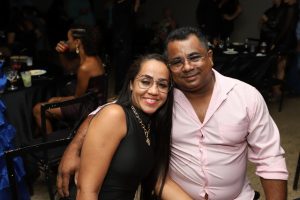 Portal Jaciara Barros – Uma mulher de preto e um soldado de camisa rosa clara posam juntos e sorriem durante evento realizado em ambiente fechado. – Portal Jaciara Barros