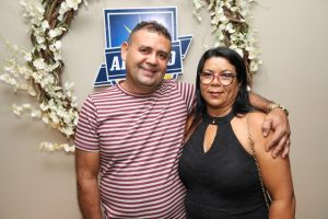 Portal Jaciara Barros – Um homem com uma camisa listrada e uma mulher com um vestido preto sorriem juntos em frente a um cenário floral. – Portal Jaciara Barros