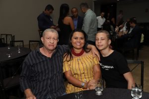 Portal Jaciara Barros – Três pessoas sentam-se à mesa com bebidas e posam ao lado de um soldado durante confraternização em local fechado. – Portal Jaciara Barros