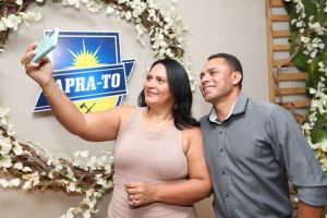 Portal Jaciara Barros – Duas pessoas tiram selfie sorrindo diante de uma coroa de flores e placa azul da APRA-TO na Praça Soldado. – Portal Jaciara Barros