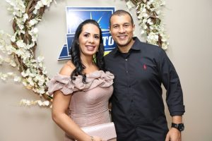 Portal Jaciara Barros – Uma mulher de vestido rosa e um homem com camisa preta, trajando estilo militar, posam sorrindo ao lado de uma coroa floral e placa azul. – Portal Jaciara Barros