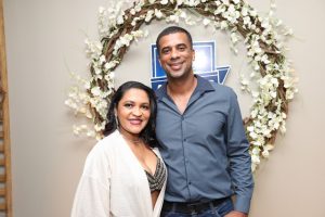 Portal Jaciara Barros – Um homem e uma mulher sorriem em frente a uma guirlanda de flores e a uma placa azul e branca com os dizeres "soldado" em parede bege. – Portal Jaciara Barros