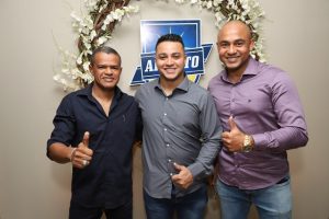 Portal Jaciara Barros – Três homens posam sorrindo e fazendo sinal de positivo diante de um painel floral em ambiente interno. – Portal Jaciara Barros